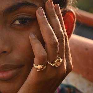 May include: Two gold rings on a person's finger. One ring features a snake design, while the other ring has a circular design with a textured surface.