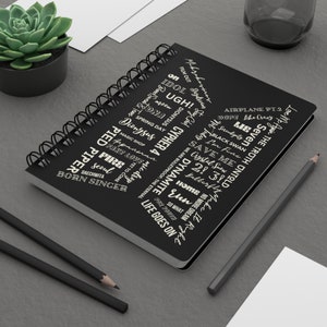 Può includere: Un quaderno nero a spirale con testo bianco. Il testo include i titoli delle canzoni della boy band sudcoreana BTS, tra cui "Dynamite", "Life Goes On", "Fake Love", "Home", "Idol", "Spring Day", "Airplane Pt.2", "Save Me", "Lie", "Black Swan", "No More Dream", "Not Today", "Serendipity", "Crystal Snow", "Butterflies", "Ddaechwita", "Born Singer", "Magic Shop", "Pied Piper", "Yet To Come", "Run", "So What", "Euphoria", "Cypher 4", "Mic Drop", "We Are Bulletproof The Eternal", "Mikrokosmos", "Film Out", "Daydream", "Love Me Again" e "The Truth Untold".