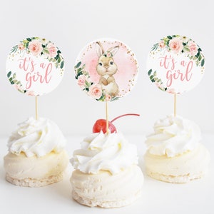 May include: Three cupcakes with white frosting and a pink and green floral design cupcake topper with the text "it's a girl". The center cupcake has a bunny rabbit topper.