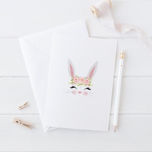May include: A white bunny with pink eyes and a pink flower crown. The bunny is on a white card with a white background.