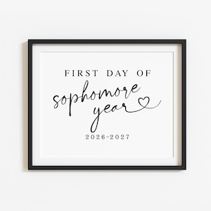 May include: A black rectangular frame containing a white sign with black text that reads FIRST DAY OF sophomore year followed by a heart symbol and the dates 2026-2027. The text uses a combination of block lettering and cursive script.