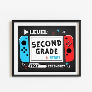 May include: A framed print featuring a black chalkboard background with a video game controller theme. The text reads LEVEL: SECOND GRADE, START, and LOADING... 2026-2027. It includes blue and red controller side panels with buttons.