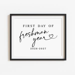 May include: A black rectangular frame containing a white sign with black text that reads FIRST DAY OF freshman year followed by a small heart symbol and the academic dates 2026-2027 printed in a combination of serif and script fonts.