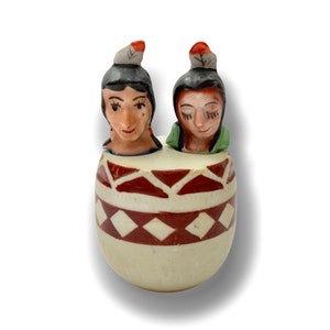 Puede incluir: Un juego de salero y pimentero de cerámica con dos figuras de nativos americanos con tocados de plumas. Las figuras están pintadas en marrón, blanco y rojo, y están sentadas dentro de un recipiente de cerámica con un patrón geométrico marrón y blanco.