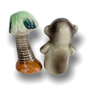 May include: A set of ceramic salt and pepper shakers. The salt shaker is shaped like a palm tree with green leaves and a brown trunk. The pepper shaker is shaped like a monkey with a brown and grey body.