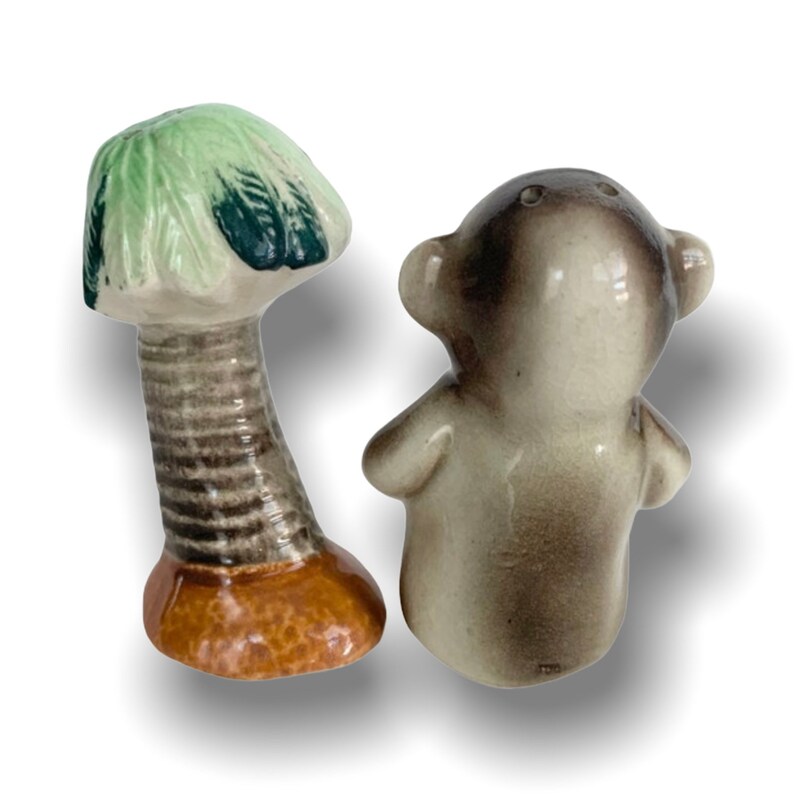 May include: A set of ceramic salt and pepper shakers. The salt shaker is shaped like a palm tree with green leaves and a brown trunk. The pepper shaker is shaped like a monkey with a brown and grey body.