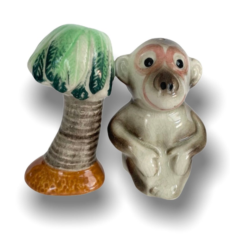 May include: A ceramic salt and pepper shaker set featuring a monkey and a palm tree. The monkey is sitting with its hands clasped in front of it. The palm tree has a green top and a brown trunk.