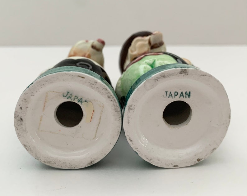 May include: A pair of vintage salt and pepper shakers depicting a man and woman in traditional clothing. The shakers are made of ceramic and have a green, white, and black color scheme. The bottom of each shaker is marked "JAPAN".