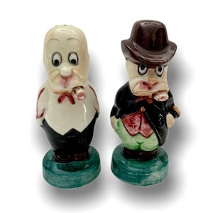 May include: A set of two ceramic salt and pepper shakers. The shakers are shaped like cartoon men with exaggerated features. One shaker has a white shirt and black pants, while the other has a brown hat, red vest, and green pants. Both shakers have a green base.
