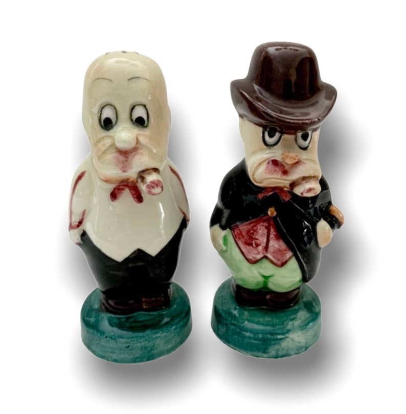 May include: A set of two ceramic salt and pepper shakers. The shakers are shaped like cartoon men with exaggerated features. One shaker has a white shirt and black pants, while the other has a brown hat, red vest, and green pants. Both shakers have a green base.