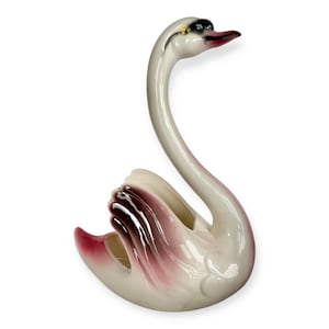 May include: A ceramic swan ornament in white, with a long, curved neck and a graceful pose. The swan's wings and beak are accented with shades of burgundy and pink. The figurine is a decorative item.