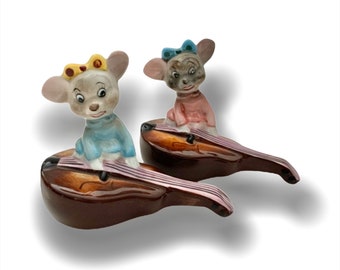 May include: Two ceramic figurines of mice playing the violin. The mice are wearing blue and pink dresses with yellow and blue bows. The violins are brown with a reddish-brown finish.