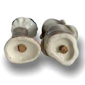 May include: A pair of ceramic salt and pepper shakers shaped like a seated couple. The shakers are painted in shades of grey and white, with a brown base. The shakers have cork stoppers on the bottom.