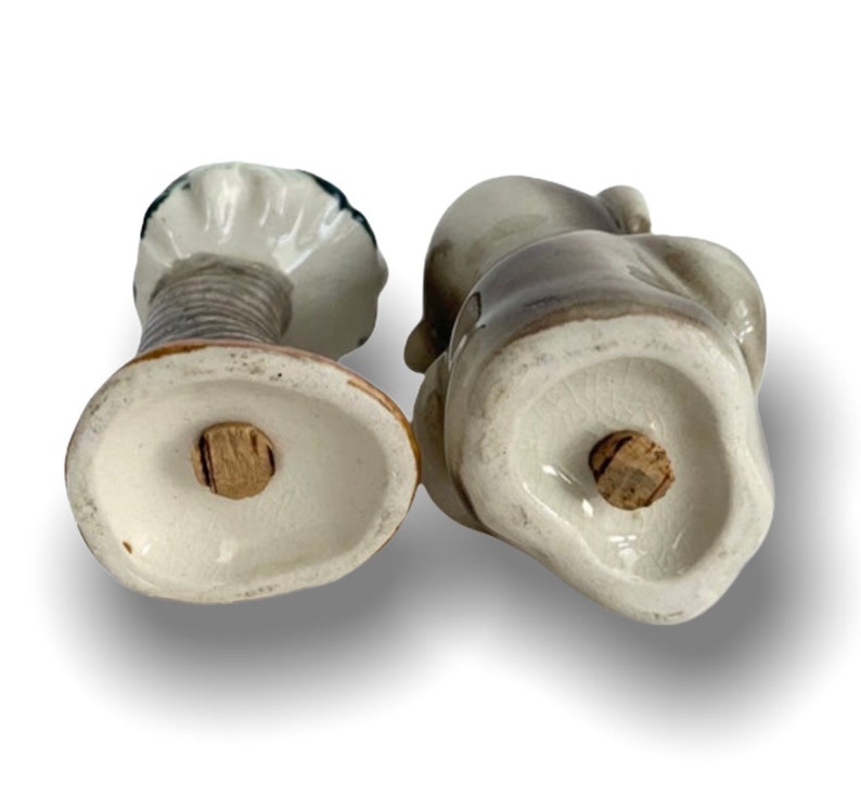 May include: A pair of ceramic salt and pepper shakers shaped like a seated couple. The shakers are painted in shades of grey and white, with a brown base. The shakers have cork stoppers on the bottom.