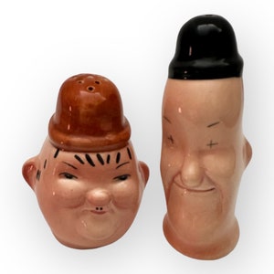 May include: A pair of ceramic salt and pepper shakers shaped like cartoon faces. The salt shaker is a brown-haired man with a brown hat and a mischievous smile. The pepper shaker is a man with black hair and a black hat, with a grumpy expression.