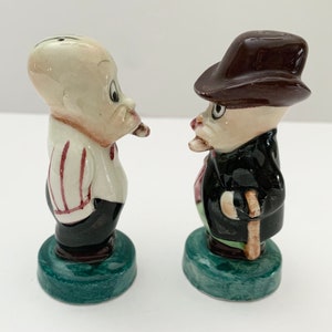 May include: A set of two ceramic salt and pepper shakers. The shakers are shaped like cartoon men. One man is wearing a striped shirt and a black hat. The other man is wearing a black suit and a brown hat. Both men have a mustache and are holding a pipe.