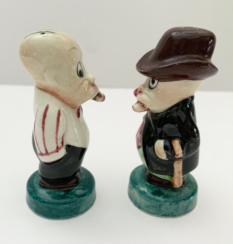May include: A set of two ceramic salt and pepper shakers. The shakers are shaped like cartoon men. One man is wearing a striped shirt and a black hat. The other man is wearing a black suit and a brown hat. Both men have a mustache and are holding a pipe.