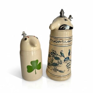 May include: Two cream-colored ceramic steins with lids. The shorter stein features a green shamrock design. The taller stein is decorated with blue and green floral patterns, gold trim, and a metallic lid topper shaped like a bird figure.