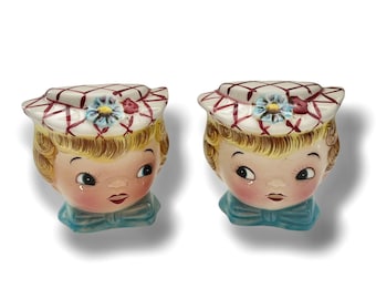 May include: A pair of vintage ceramic salt and pepper shakers shaped like children's heads. The shakers are painted with a white and red plaid hat, blonde hair, and a blue bow tie.