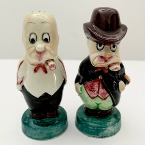 May include: A pair of vintage ceramic salt and pepper shakers. The shakers are shaped like men with exaggerated features, wearing suits and hats. One shaker has a white shirt and a red bow tie, while the other has a green shirt and a brown hat. Both shakers have a green base.