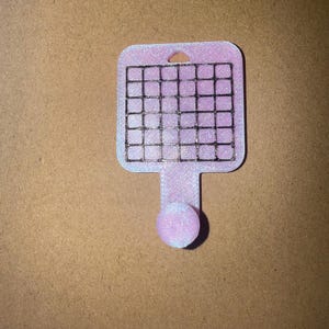 May include: A pink and white 3D-printed kitchen tool with a grid of wires. The tool has a handle with a small ball at the end and a hole at the top. The grid is made of black wires.