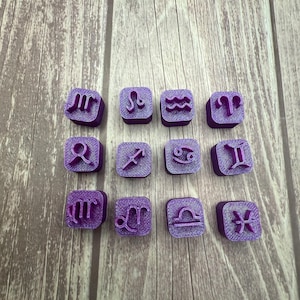 May include: A collection of twelve purple square stamps, each featuring a different zodiac symbol. The stamps are arranged in three rows of four, with each symbol clearly visible. The background is a light brown wooden surface.