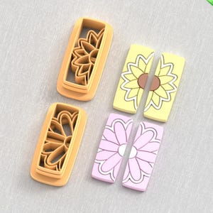 May include: A set of four items: two orange plastic sunflower-shaped cookie cutters and two rectangular flower-shaped decorations, one yellow with a brown center and one pink with a white center. The items are arranged on a light gray surface.