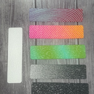 May include: Five nail files with different textures and colors. The files are arranged vertically on a wooden surface. The top file is a dark red with a swirl pattern, the second file is pink and orange with diagonal stripes, the third file is green and blue with a gradient, the fourth file is black and white with a dotted pattern, and the bottom file is black with a speckled pattern.