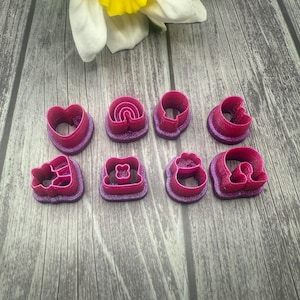 May include: A set of eight pink plastic cookie cutters in various shapes, including a heart, a rainbow, a circle, a flower, a star, a square, a cloud, and a bell. The cutters are arranged on a wooden surface.