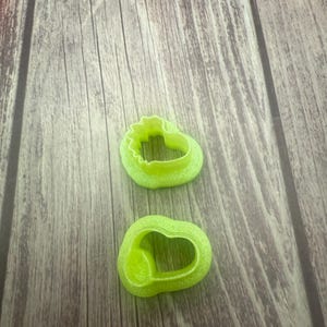 May include: Two green plastic cookie cutters in the shape of hearts. The cutters are on a wooden surface.