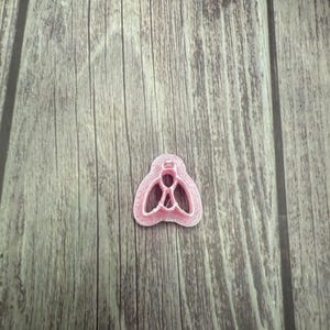 May include: A pink plastic cookie cutter in the shape of a fly. The cutter has a detailed design with cut-out sections for the wings and body. The cutter is on a wooden surface.