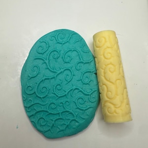 May include: A teal-colored, oval-shaped clay piece with a swirling vine pattern, next to a yellow cylindrical rolling pin with a matching design. The tools are likely for crafting or baking, featuring a detailed, embossed texture.