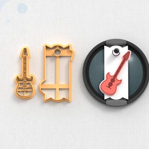 May include: Three items related to music. Two are gold-colored cookie cutters in the shape of a bass guitar and a rectangular frame. The third is a red bass guitar on a white tag, set against a black and gray background.