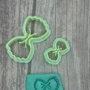 May include: Three light green bow-shaped cookie cutters in different sizes, along with a teal-colored bow-shaped imprint. The cutters have a scalloped edge and are made of plastic. The items are displayed on a wooden surface.