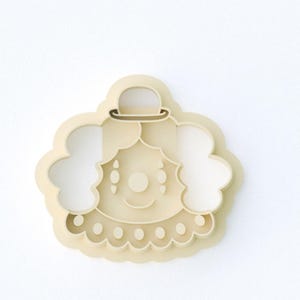 May include: A beige plastic cookie cutter in the shape of a smiling clown face with a small hat. The clown has a round nose, teardrop-shaped eyes, and a scalloped collar. The cutter is designed for baking.