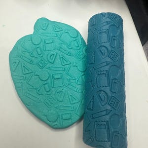 May include: A teal heart-shaped clay piece and a blue cylindrical clay roller, both embossed with school-themed designs. The designs include pencils, protractors, notebooks, and other school supplies. The items are on a white surface.