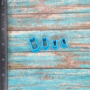 May include: Four blue plastic cookie cutters shaped like bunnies, arranged in a row from largest to smallest on a light blue wooden surface.