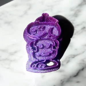 May include: Purple 3D printed cookie cutter shaped like a cartoon character with a hat. The character has a round head, large eyes, and a small mouth.
