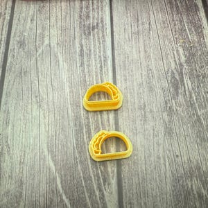May include: Two yellow plastic cookie cutters shaped like tacos. The cutters are on a wooden surface.