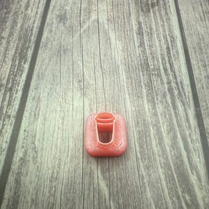 May include: A pink plastic cookie cutter shaped like a cup with a square frame.