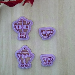 May include: Four light purple plastic cookie cutters featuring skull-themed designs. Two cutters are shaped like teapots with skull faces, and two are shaped like teacups with skull faces. The items have a textured, 3D-printed appearance.