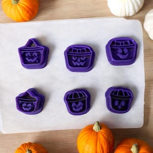 May include: Six purple Halloween-themed cookie cutters in various shapes, including jack-o'-lantern buckets. The cutters are arranged on a white surface, with orange pumpkins and white gourds in the background. Perfect for making festive treats.