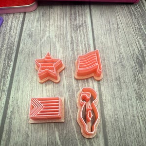May include: Four red plastic cookie cutters in the shapes of a star, a flag, a striped rectangle, and a bell. The cutters are on a wooden surface.
