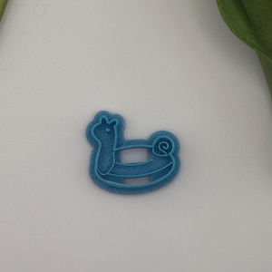 May include: A blue plastic cookie cutter in the shape of a rocking horse. The cutter has a detailed design, including the horse's head, body, and curved rockers. The cutter is designed for making cookies or other treats.