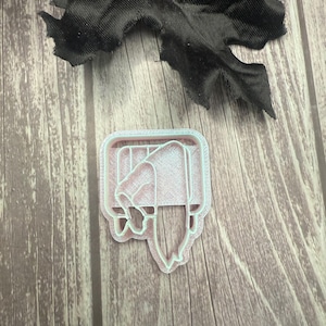 May include: A light pink plastic cookie cutter in the shape of a hand holding a knife. The cutter has a rectangular frame with a detailed design. A black fabric flower is in the background.