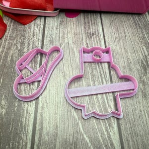 May include: Two pink plastic cookie cutters, one shaped like a hat and the other shaped like a sandal. The cutters are on a wooden surface.