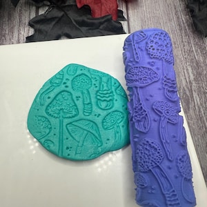 May include: A teal clay piece with mushroom imprints next to a purple roller with mushroom designs. The roller is cylindrical and features various mushroom shapes. The clay piece is a flattened oval shape with detailed mushroom impressions.