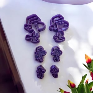 May include: Set of six purple plastic cookie cutters shaped like Snoopy wearing a Santa hat. The cutters are in two sizes, with the larger cutters featuring Snoopy standing and the smaller cutters featuring Snoopy sitting.