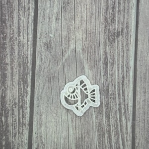 May include: A silver-colored fish-shaped cookie cutter with intricate cut-out details. The cutter features a stylized design of a fish, including eyes, fins, and scales. The cutter is placed on a wooden surface.
