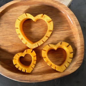 May include: Three heart-shaped cookie cutters made of yellow plastic, arranged on a wooden surface.
